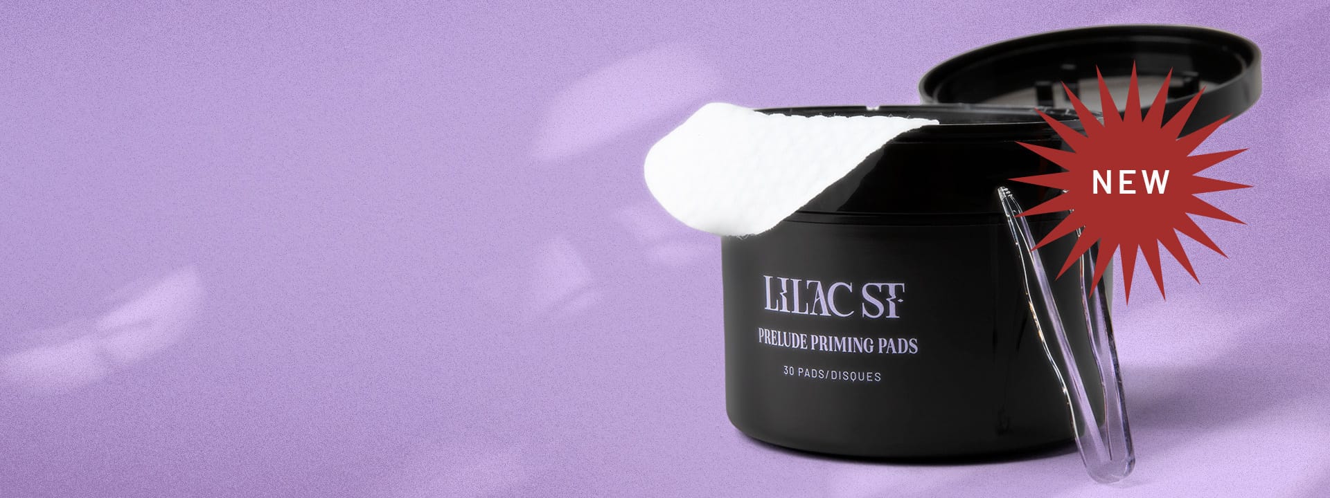 NEW: An open canister of Lilac St. Prelude Priming Pads showing one of the white circular pads draped over the open lip of the container and a pair of clear tweezers leaning tip-up on the container on top of a seamless lilac colored background dappled with streaks of light. 