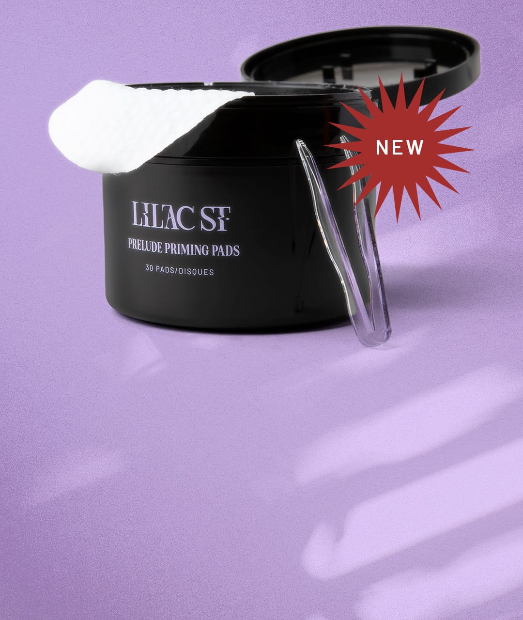 NEW: An open canister of Lilac St. Prelude Priming Pads showing one of the white circular pads draped over the open lip of the container and a pair of clear tweezers leaning tip-up on the container on top of a seamless lilac colored background dappled with streaks of light. 